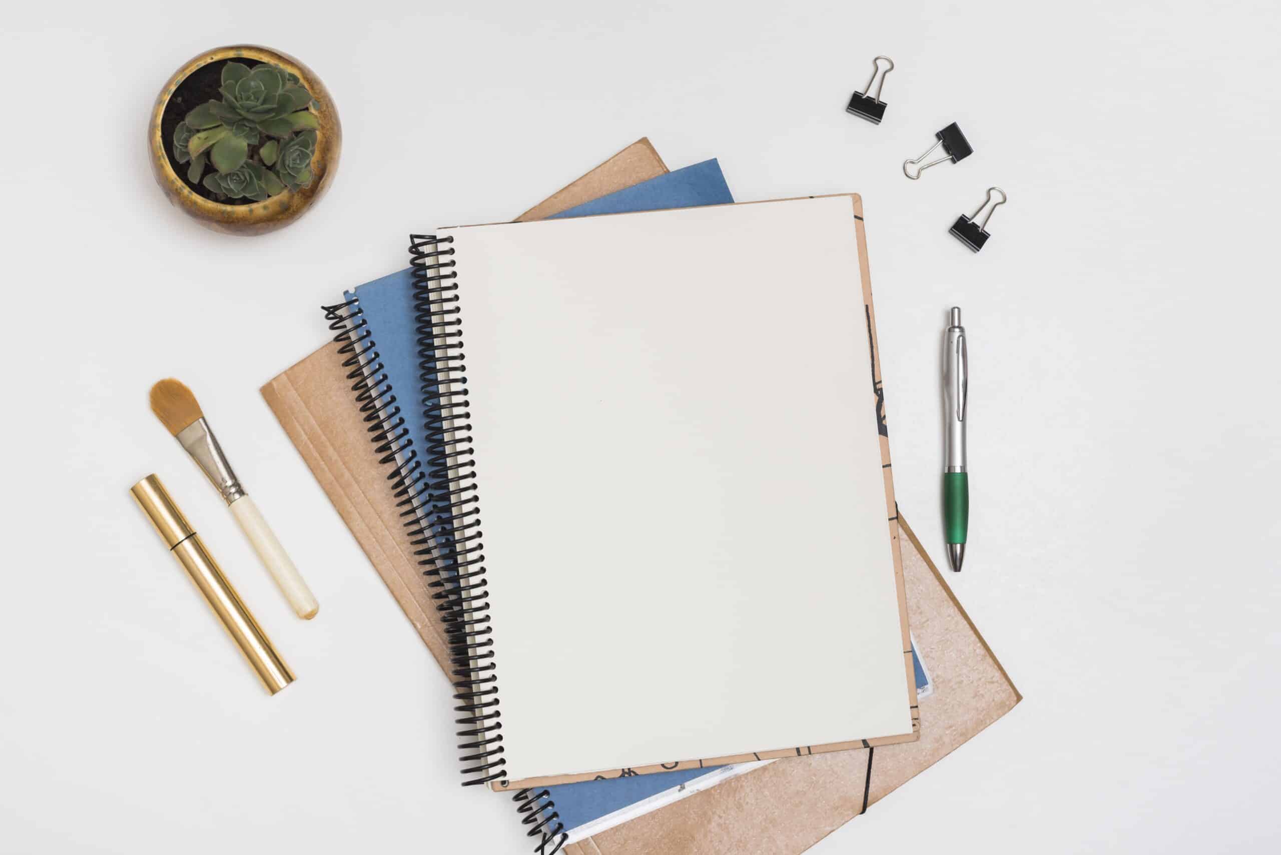blank-spiral-note-with-make-up-brush-mascara-paper-clips-pen-with-potted-plant-white-desk