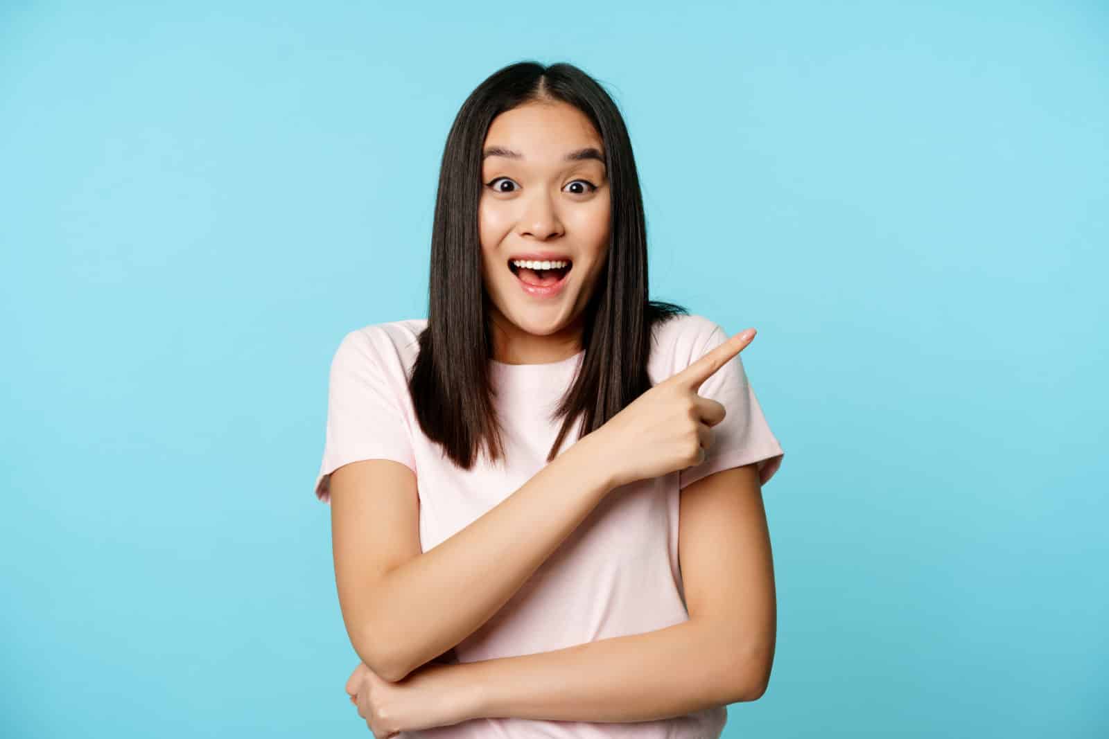happy-asian-woman-staring-surprised-pointing-at-upper-right-corner-showing-fantastic-discount-sale-advertisement-blue-background